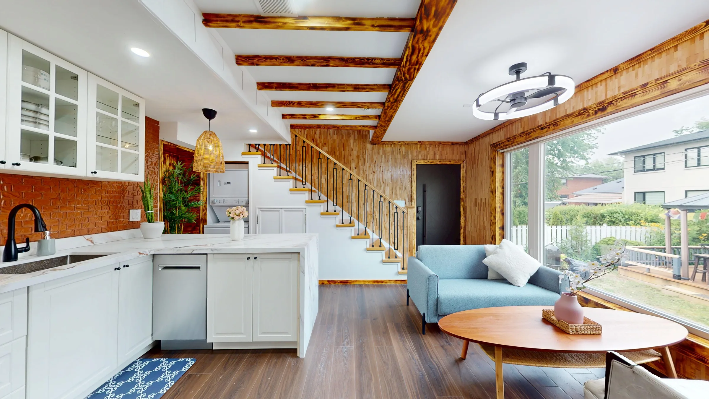 Open-plan kitchen and living room with exposed burnt-wood beams, a floating staircase, and a wall of windows onto the backyard