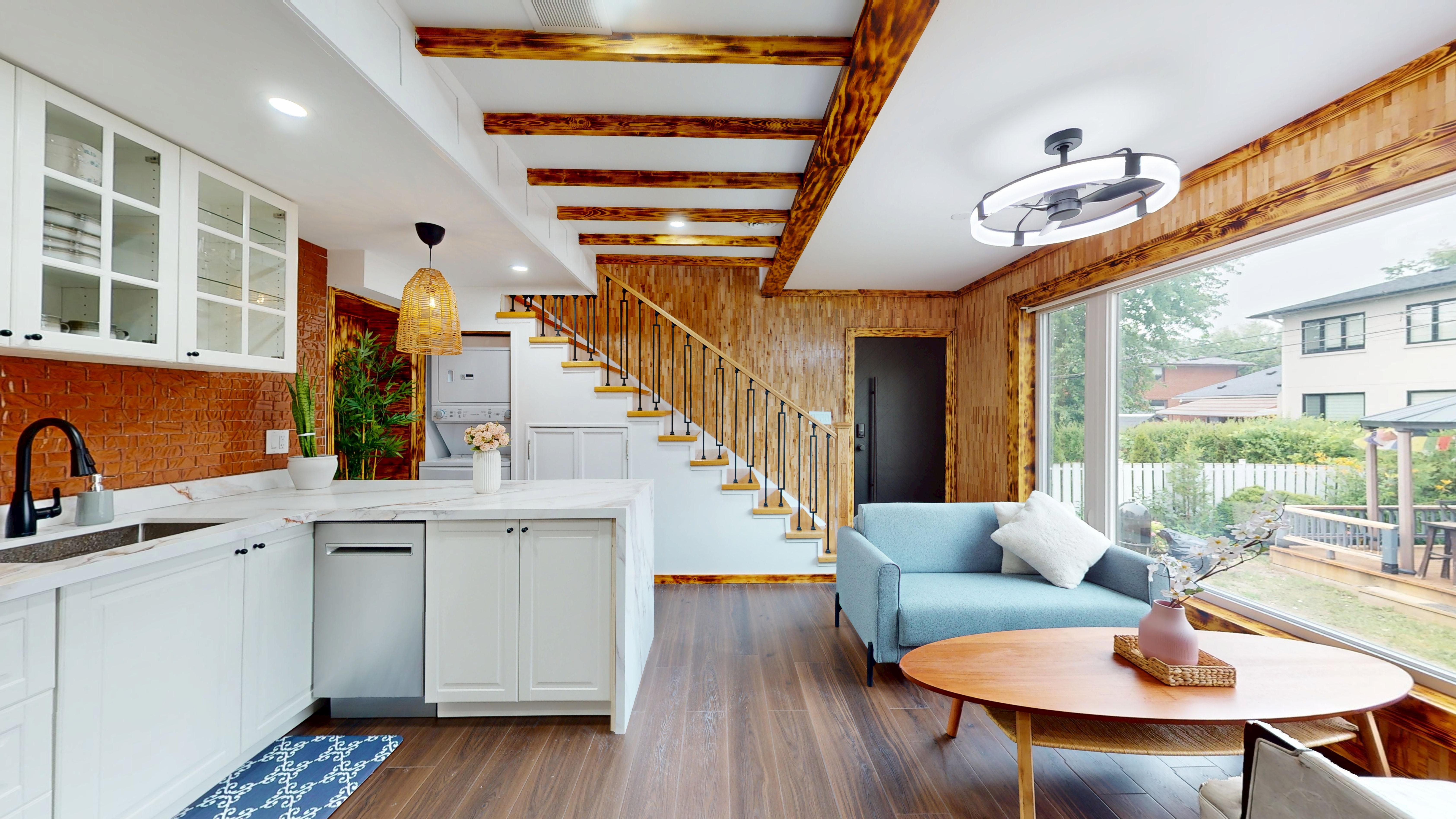 Open-plan kitchen and living room with exposed burnt-wood beams, a floating staircase, and a wall of windows onto the backyard
